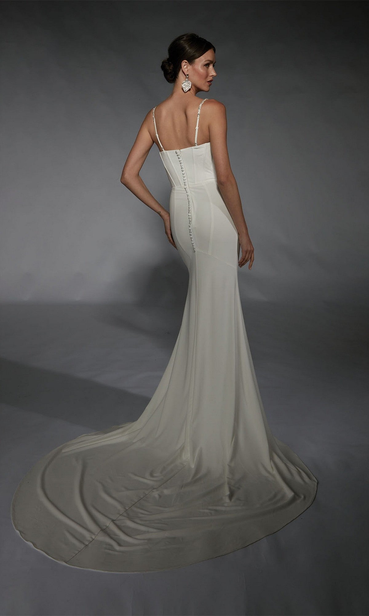 A woman stands facing away in the Alyce 7103, a fitted long white dress with thin straps, a long train, and button details down the back, highlighting the gown’s elegant formal style.